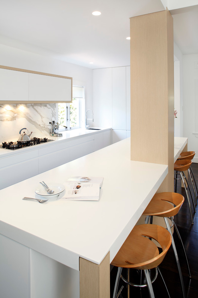 Example of a minimalist single-wall eat-in kitchen design in Sydney with a drop-in sink, flat-panel cabinets, white cabinets, solid surface countertops, stone slab backsplash and stainless steel appliances