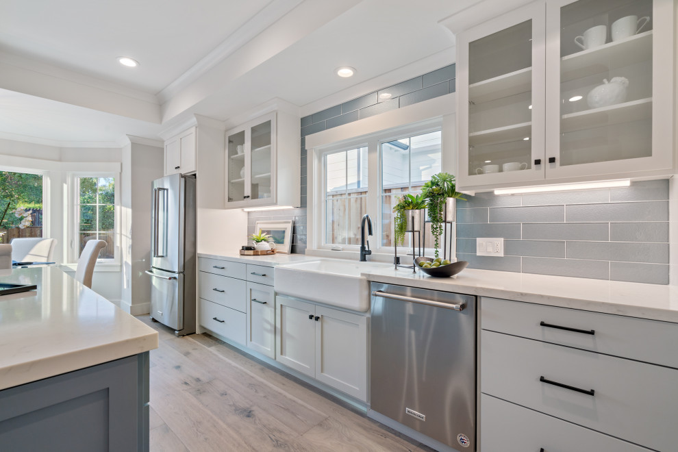 Bild på ett mellanstort amerikanskt vit vitt kök, med en rustik diskho, skåp i shakerstil, vita skåp, bänkskiva i kvarts, blått stänkskydd, stänkskydd i glaskakel, rostfria vitvaror, ljust trägolv, en köksö och grått golv
