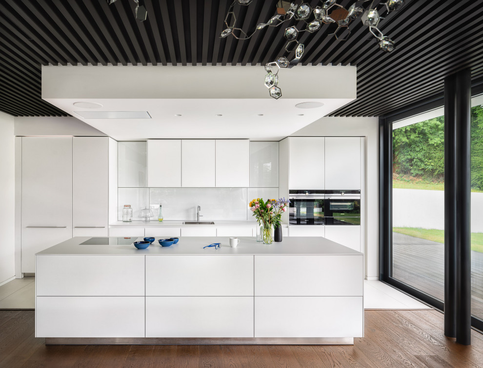 Design ideas for a large contemporary galley open plan kitchen in London with a submerged sink, flat-panel cabinets, white cabinets, white splashback, glass sheet splashback, integrated appliances, medium hardwood flooring, an island, beige floors and white worktops.