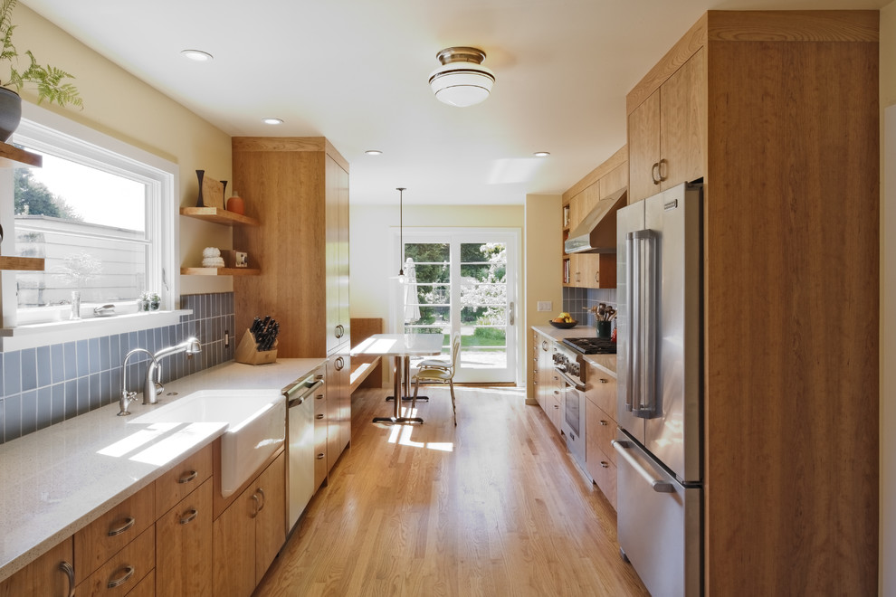Transitional galley enclosed kitchen photo in Portland with a farmhouse sink, stainless steel appliances, flat-panel cabinets, medium tone wood cabinets, recycled glass countertops, blue backsplash and ceramic backsplash