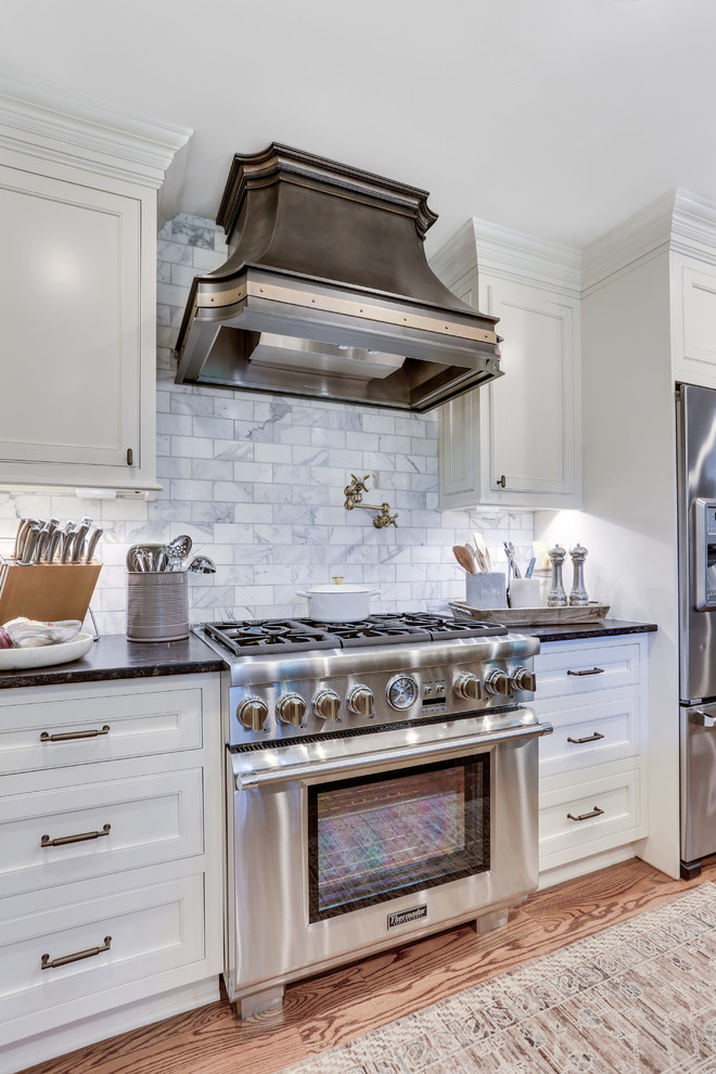 This is an example of a medium sized traditional u-shaped kitchen/diner in DC Metro with a belfast sink, beaded cabinets, white cabinets, quartz worktops, grey splashback, marble splashback, stainless steel appliances, light hardwood flooring, an island and brown floors.
