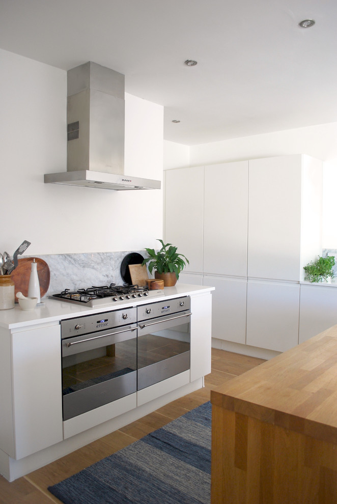 Design ideas for a large modern l-shaped kitchen/diner in Wiltshire with a single-bowl sink, flat-panel cabinets, white cabinets, wood worktops, grey splashback, stone tiled splashback, stainless steel appliances, light hardwood flooring and an island.