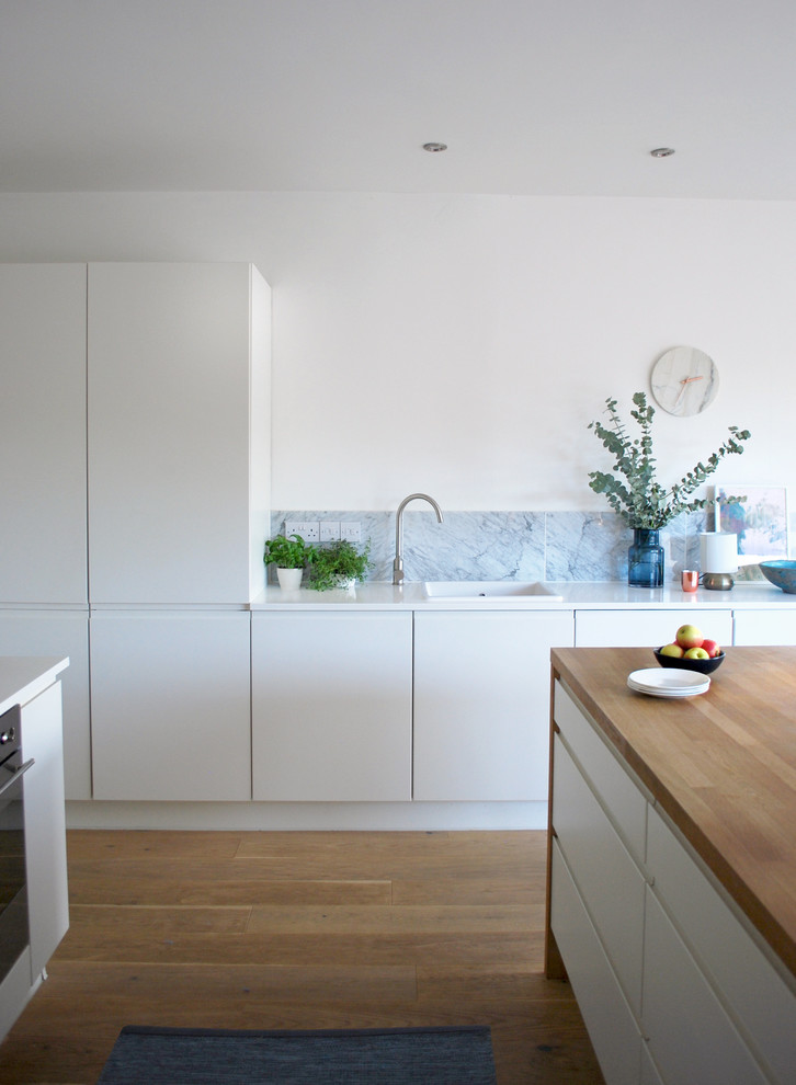 Modern inredning av ett stort kök, med en enkel diskho, släta luckor, vita skåp, träbänkskiva, grått stänkskydd, stänkskydd i stenkakel, rostfria vitvaror, ljust trägolv och en köksö