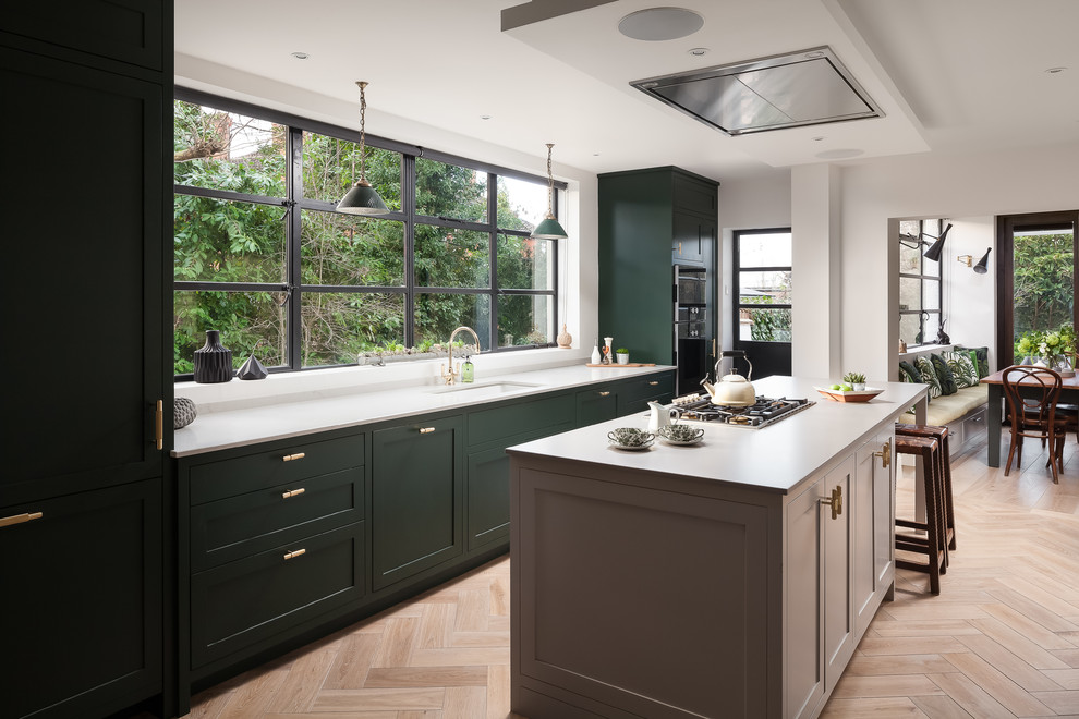 На фото: параллельная кухня-гостиная в стиле неоклассика (современная классика) с тройной мойкой, фасадами в стиле шейкер, зелеными фасадами, черной техникой, светлым паркетным полом и островом