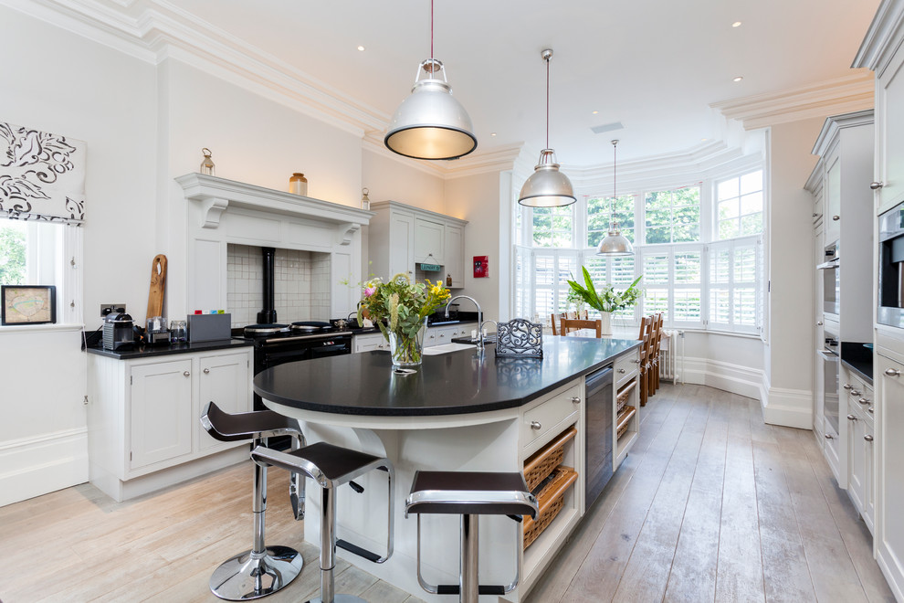 Photo of a classic galley kitchen/diner in London with a belfast sink, shaker cabinets, grey cabinets, white splashback, ceramic splashback, light hardwood flooring and an island.