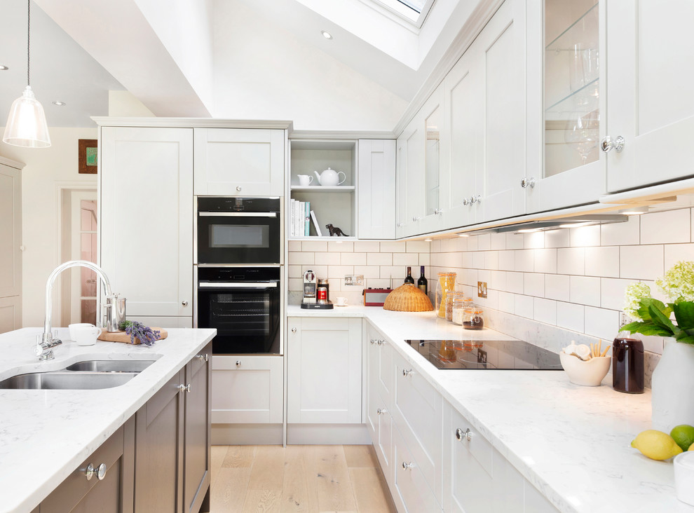 Photo of a medium sized traditional l-shaped kitchen/diner in London with a built-in sink, shaker cabinets, grey cabinets, engineered stone countertops, white splashback, porcelain splashback, black appliances, light hardwood flooring, an island and white worktops.