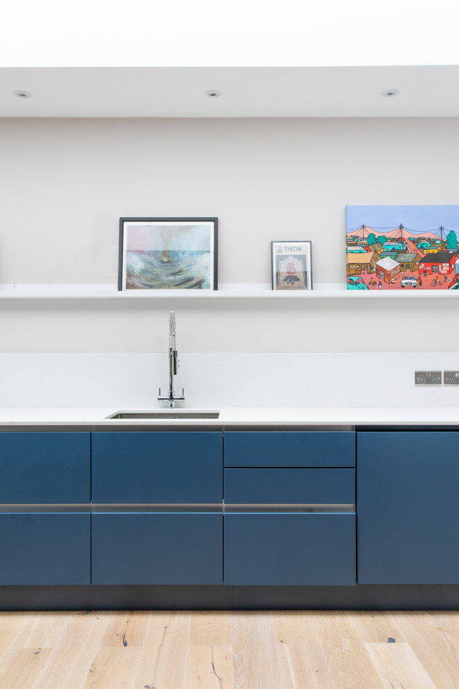 Mittelgroße Moderne Wohnküche mit Waschbecken, flächenbündigen Schrankfronten, blauen Schränken, Mineralwerkstoff-Arbeitsplatte, Küchenrückwand in Weiß, Elektrogeräten mit Frontblende, hellem Holzboden, Kücheninsel und weißer Arbeitsplatte in London