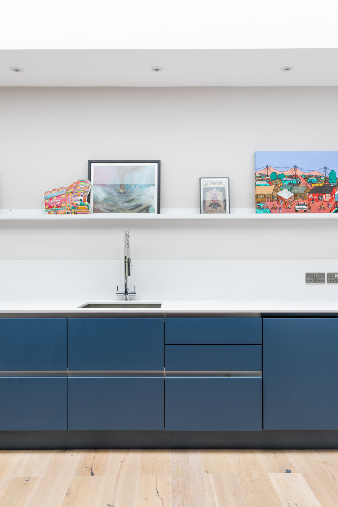 Photo of a medium sized contemporary kitchen/diner in London with a single-bowl sink, flat-panel cabinets, blue cabinets, composite countertops, white splashback, integrated appliances, light hardwood flooring, an island and white worktops.