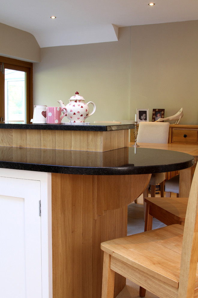 Expansive contemporary u-shaped open plan kitchen in Hampshire with a belfast sink, shaker cabinets, white cabinets, granite worktops, black splashback, black appliances, limestone flooring and a breakfast bar.
