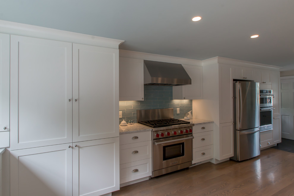 Example of a classic u-shaped medium tone wood floor kitchen design in Boston with a farmhouse sink, recessed-panel cabinets, white cabinets, quartz countertops, blue backsplash, glass tile backsplash, stainless steel appliances and an island