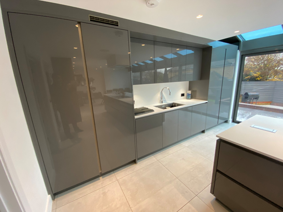 Photo of a large contemporary l-shaped kitchen/diner in London with a built-in sink, flat-panel cabinets, grey cabinets, quartz worktops, white splashback, glass sheet splashback, black appliances, porcelain flooring, an island, grey floors, white worktops and a drop ceiling.