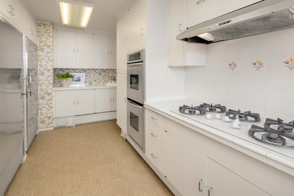 Inspiration for a large 1960s u-shaped laminate floor and brown floor eat-in kitchen remodel in Seattle with flat-panel cabinets, white cabinets, tile countertops, white backsplash, porcelain backsplash, stainless steel appliances, an island and white countertops