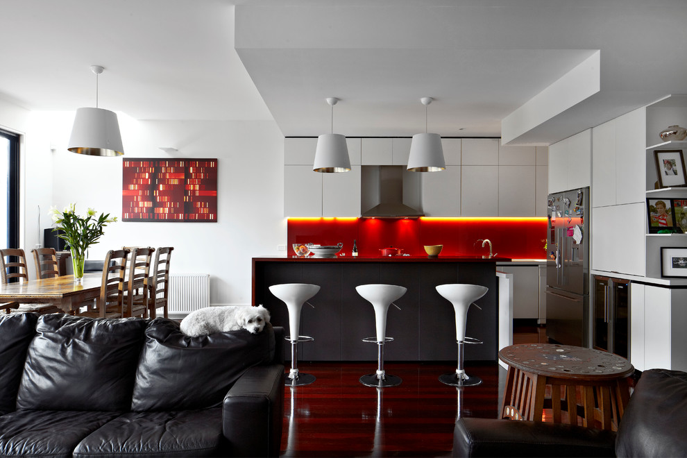 На фото: кухня-гостиная в современном стиле с плоскими фасадами, красным фартуком, техникой из нержавеющей стали, темным паркетным полом, островом и черно-белыми фасадами с
