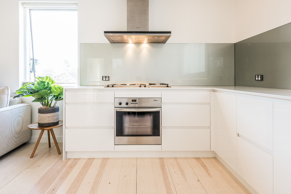 Immagine di una piccola cucina a L moderna con lavello a doppia vasca, ante bianche, paraspruzzi verde, paraspruzzi con lastra di vetro, elettrodomestici in acciaio inossidabile e parquet chiaro