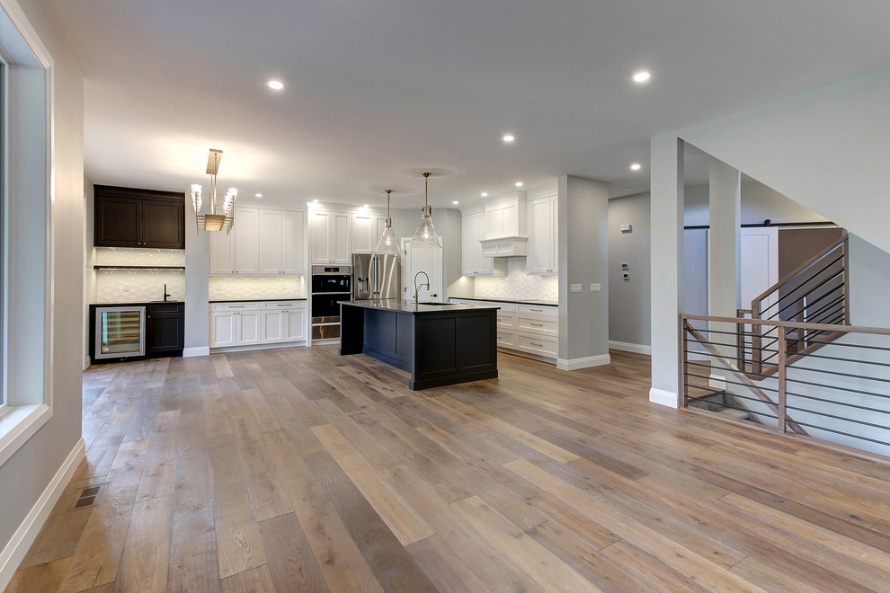 Example of a mid-sized transitional l-shaped medium tone wood floor and brown floor open concept kitchen design in Edmonton with an undermount sink, recessed-panel cabinets, white cabinets, quartz countertops, white backsplash, ceramic backsplash, stainless steel appliances, an island and black countertops