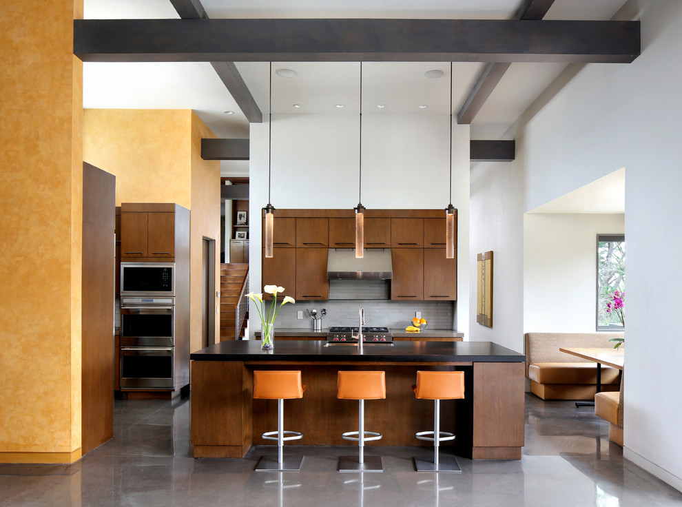 Пример оригинального дизайна: кухня в современном стиле с врезной мойкой, плоскими фасадами, темными деревянными фасадами, серым фартуком, техникой из нержавеющей стали, бетонным полом, островом и обеденным столом
