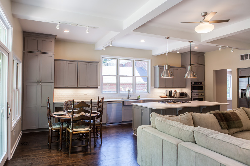 Large classic single-wall open plan kitchen in Chicago with a belfast sink, shaker cabinets, grey cabinets, engineered stone countertops, white splashback, metro tiled splashback, stainless steel appliances, dark hardwood flooring and an island.