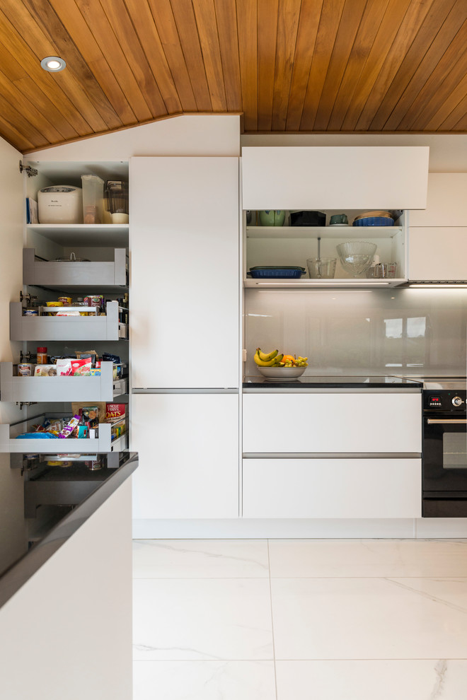 Design ideas for a medium sized contemporary galley kitchen pantry in Christchurch with a submerged sink, flat-panel cabinets, white cabinets, engineered stone countertops, grey splashback, glass sheet splashback, stainless steel appliances, ceramic flooring, an island and white floors.