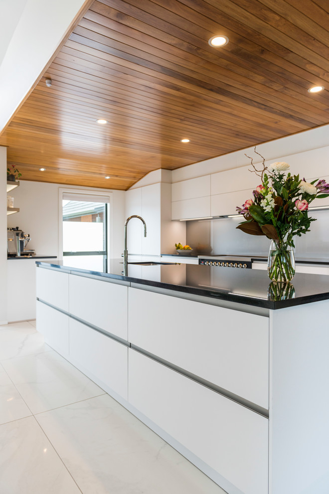 Photo of a medium sized contemporary galley kitchen pantry in Christchurch with a submerged sink, flat-panel cabinets, white cabinets, engineered stone countertops, grey splashback, glass sheet splashback, stainless steel appliances, ceramic flooring, an island and white floors.