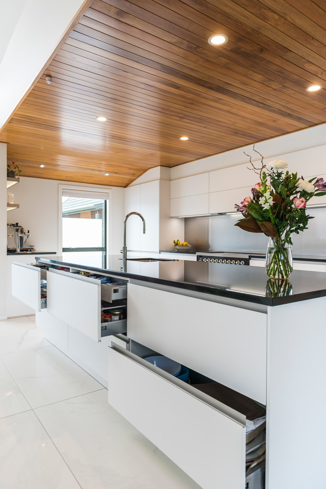 Medium sized contemporary galley kitchen pantry in Christchurch with a submerged sink, flat-panel cabinets, white cabinets, engineered stone countertops, grey splashback, glass sheet splashback, stainless steel appliances, ceramic flooring, an island and white floors.