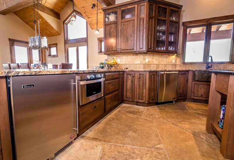 Large rustic u-shaped open plan kitchen in Denver with a belfast sink, raised-panel cabinets, medium wood cabinets, granite worktops, beige splashback, stainless steel appliances, a breakfast bar and beige floors.