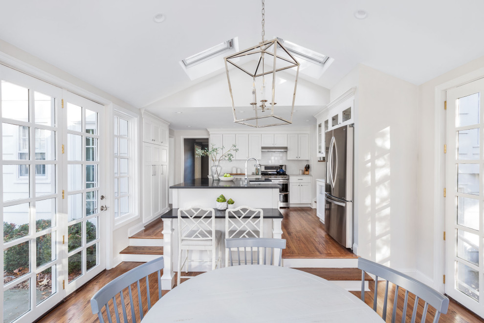 Inspiration for a coastal u-shaped medium tone wood floor, brown floor and vaulted ceiling eat-in kitchen remodel in New York with a farmhouse sink, shaker cabinets, white cabinets, white backsplash, stainless steel appliances, an island and black countertops