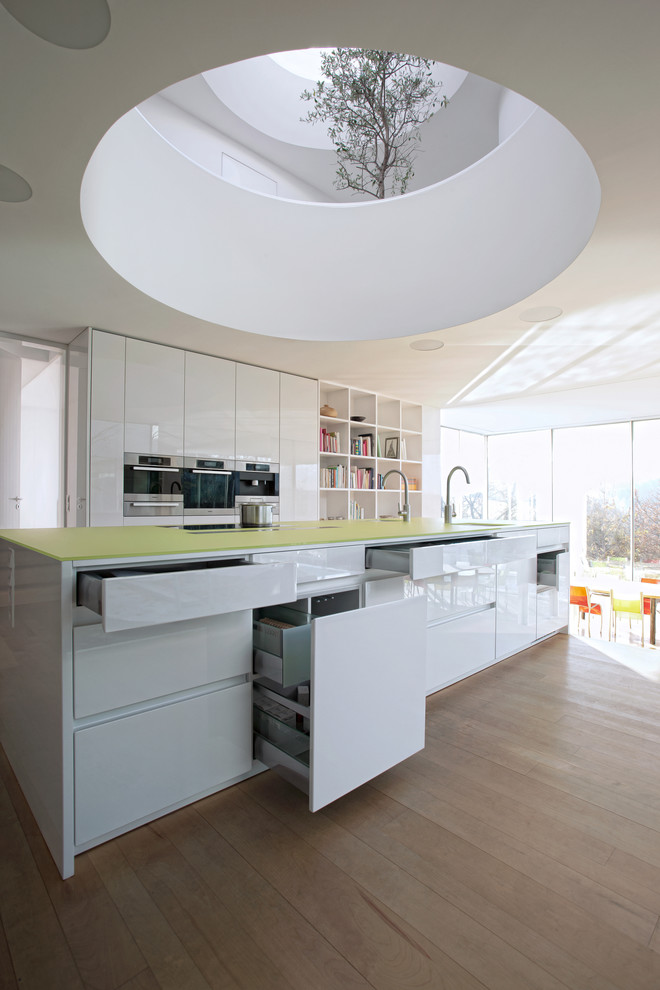 Bild på ett stort funkis kök med öppen planlösning, med släta luckor, vita skåp, bänkskiva i glas, rostfria vitvaror, mellanmörkt trägolv och en köksö