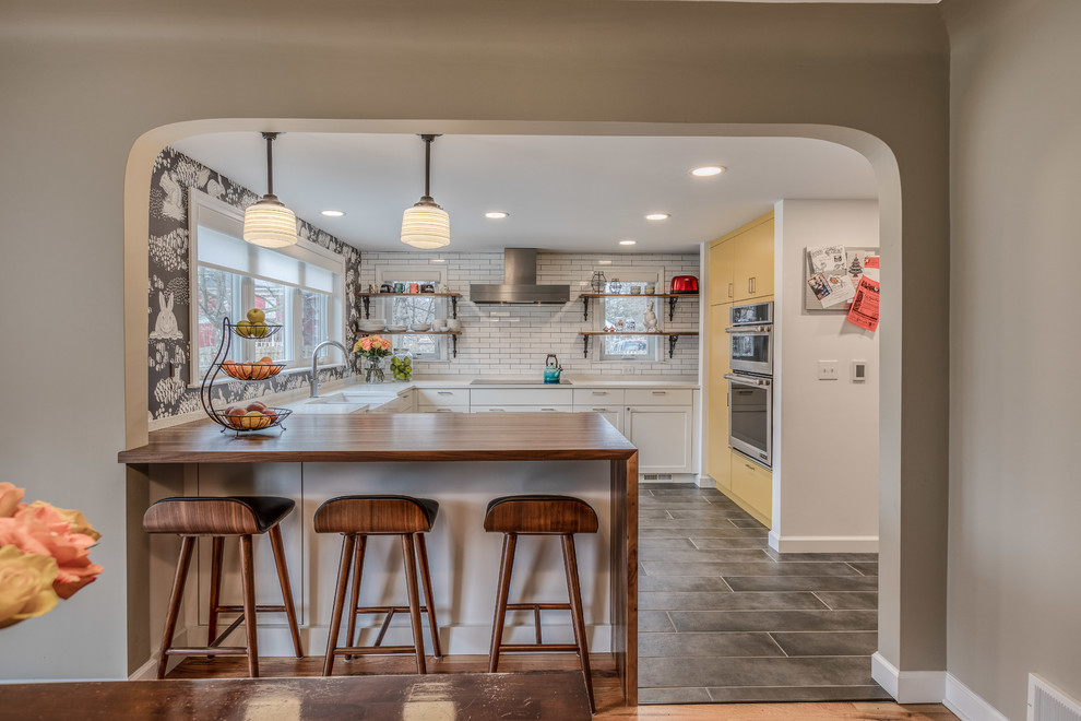 This is an example of a small rural u-shaped open plan kitchen in Detroit with a belfast sink, shaker cabinets, white cabinets, engineered stone countertops, white splashback, metro tiled splashback, stainless steel appliances, porcelain flooring, a breakfast bar, grey floors and white worktops.