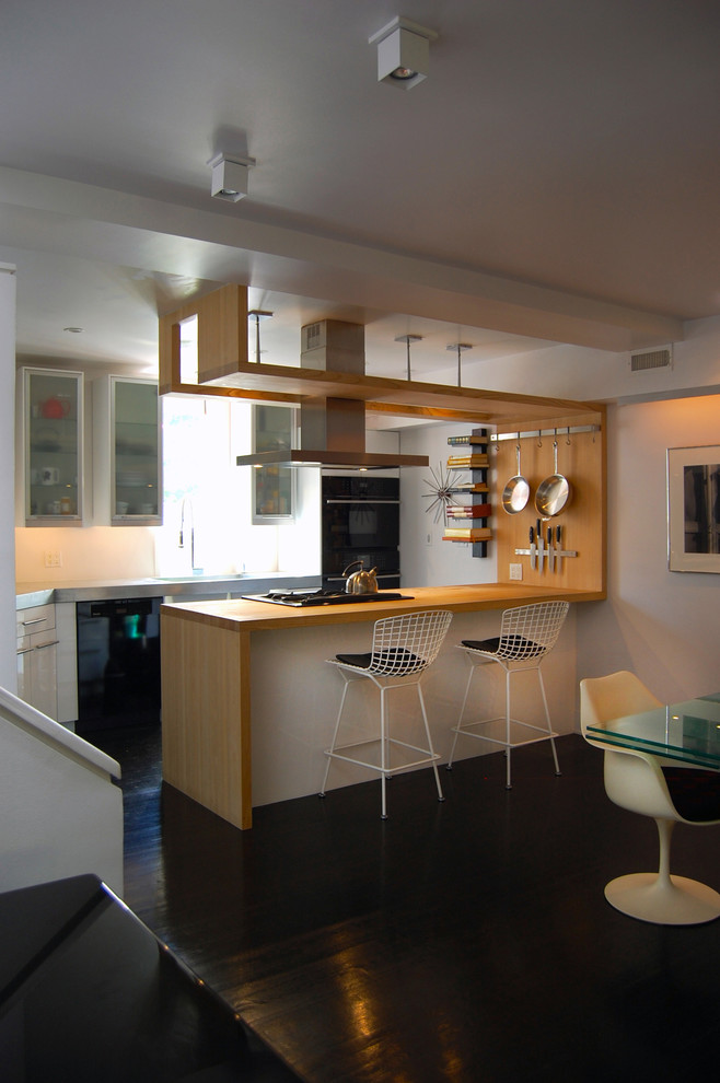Cette image montre une cuisine américaine minimaliste en U avec un placard à porte plane, des portes de placard blanches, un plan de travail en bois, un électroménager noir, parquet foncé et une péninsule.
