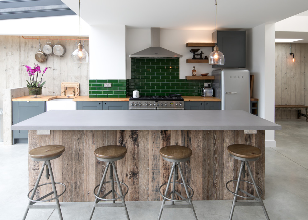 This is an example of a contemporary galley kitchen in Sussex with a belfast sink, shaker cabinets, grey cabinets, wood worktops, green splashback, metro tiled splashback, stainless steel appliances, concrete flooring, an island, grey floors and grey worktops.