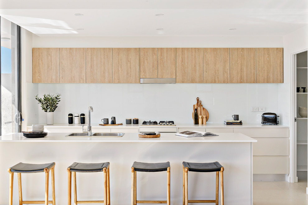 Contemporary galley kitchen in Wollongong with a built-in sink, flat-panel cabinets, white cabinets, white splashback, glass sheet splashback, integrated appliances, an island, beige floors and white worktops.