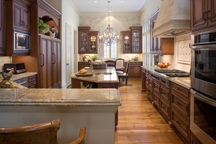 Example of a large classic galley light wood floor enclosed kitchen design in Houston with an undermount sink, raised-panel cabinets, granite countertops, beige backsplash, stainless steel appliances, medium tone wood cabinets and an island