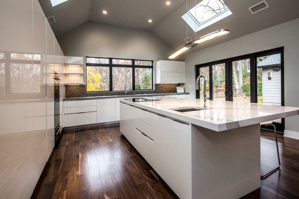 Cette image montre une cuisine américaine encastrable design avec un placard à porte plane, des portes de placard blanches, un plan de travail en quartz modifié, un sol en bois brun et îlot.