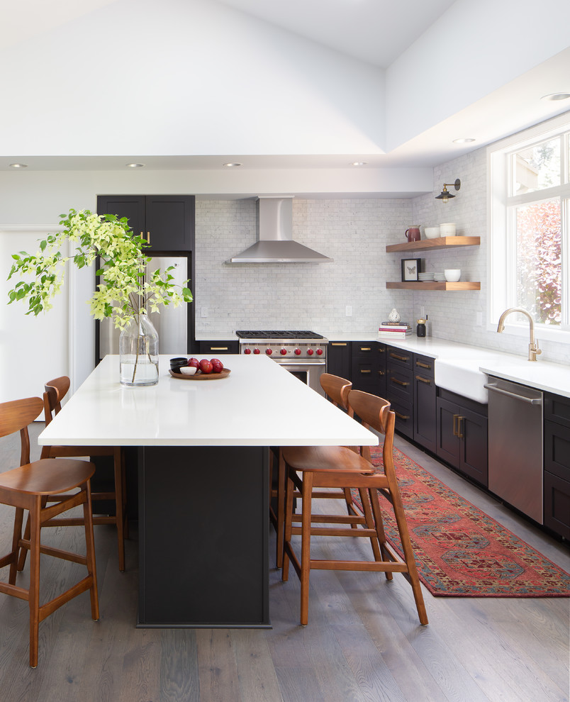 Inspiration for a transitional l-shaped medium tone wood floor and brown floor kitchen remodel in Seattle with a farmhouse sink, shaker cabinets, black cabinets, white backsplash, marble backsplash, stainless steel appliances, an island and white countertops