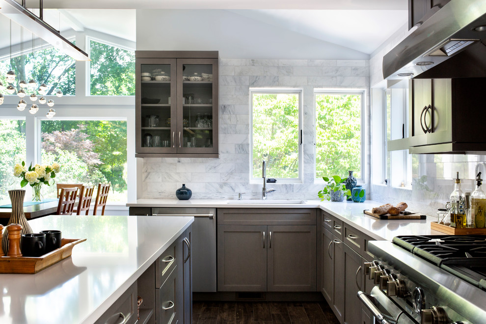 Cette photo montre une cuisine américaine chic en L avec un placard à porte shaker, des portes de placard grises, un plan de travail en surface solide, une crédence grise, îlot, un plan de travail blanc et fenêtre au-dessus de l'évier.
