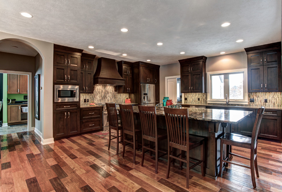 Photo of a traditional l-shaped kitchen/diner in Grand Rapids with an integrated sink, shaker cabinets, dark wood cabinets, composite countertops, brown splashback, glass tiled splashback, stainless steel appliances, medium hardwood flooring and an island.