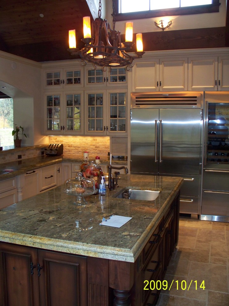 Example of a classic travertine floor kitchen design in Denver with an undermount sink, glass-front cabinets, distressed cabinets, granite countertops, multicolored backsplash, stone tile backsplash, stainless steel appliances and an island