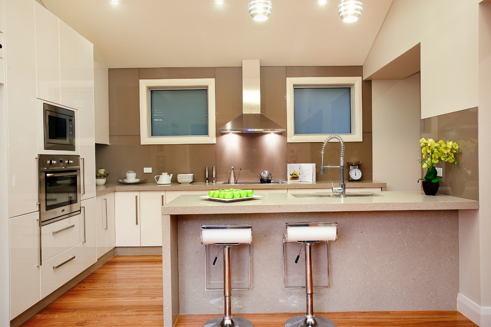 Moderne Wohnküche mit Doppelwaschbecken, flächenbündigen Schrankfronten, beigen Schränken, Küchenrückwand in Braun, Glasrückwand, braunem Holzboden, Halbinsel, Granit-Arbeitsplatte und Küchengeräten aus Edelstahl in Sydney