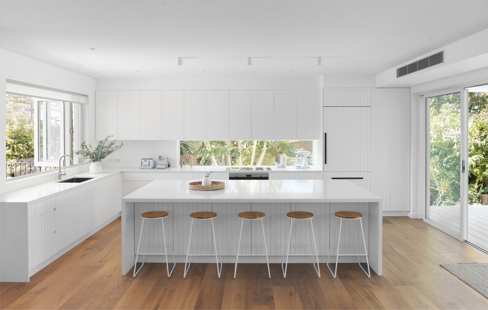 Inspiration for a coastal l-shaped kitchen in Sydney with a submerged sink, flat-panel cabinets, white cabinets, window splashback, integrated appliances, medium hardwood flooring, an island and white worktops.
