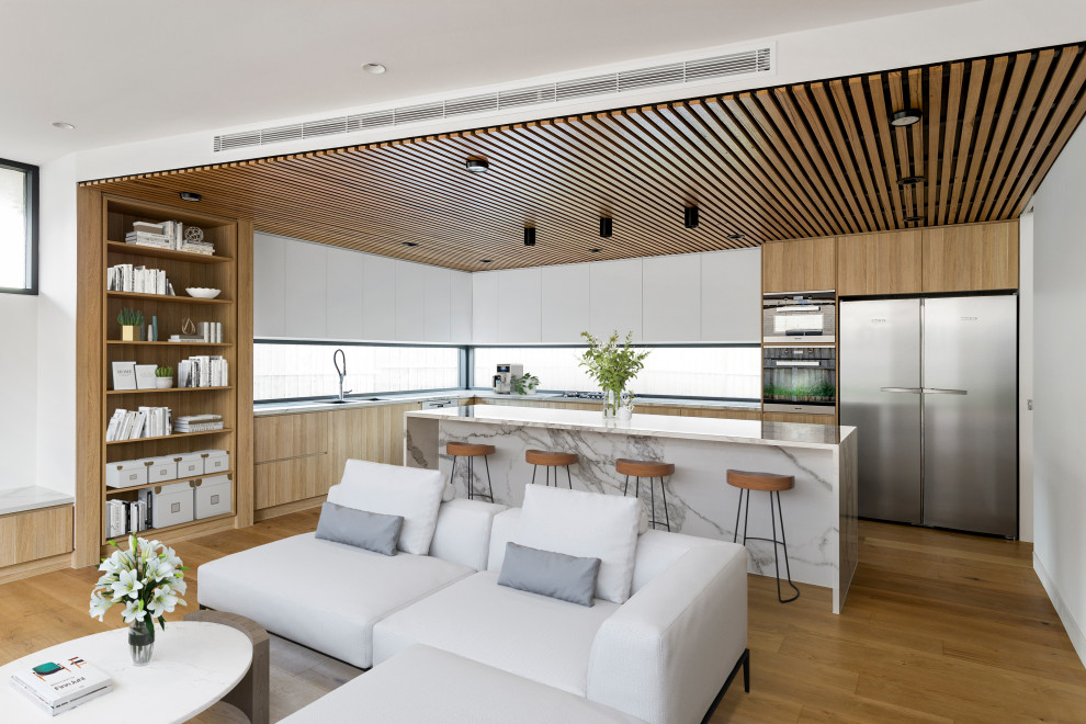 This is an example of a large contemporary l-shaped open plan kitchen in Melbourne with a submerged sink, flat-panel cabinets, light wood cabinets, marble worktops, window splashback, black appliances, light hardwood flooring, an island, brown floors, white worktops and a timber clad ceiling.
