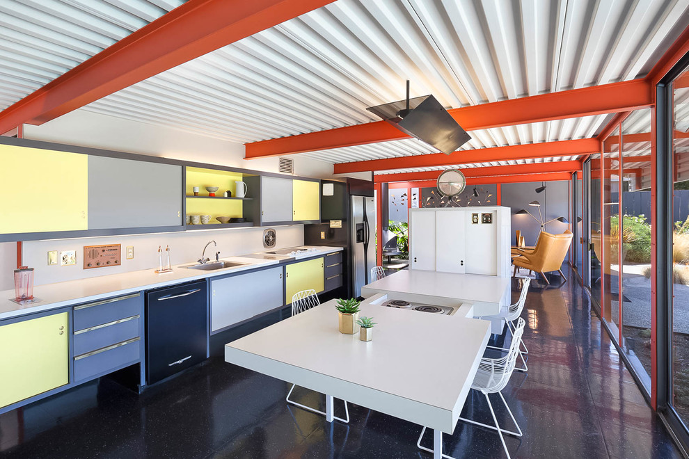 На фото: кухня-гостиная в стиле ретро с накладной мойкой, плоскими фасадами, желтыми фасадами, техникой под мебельный фасад, островом, черным полом и белой столешницей