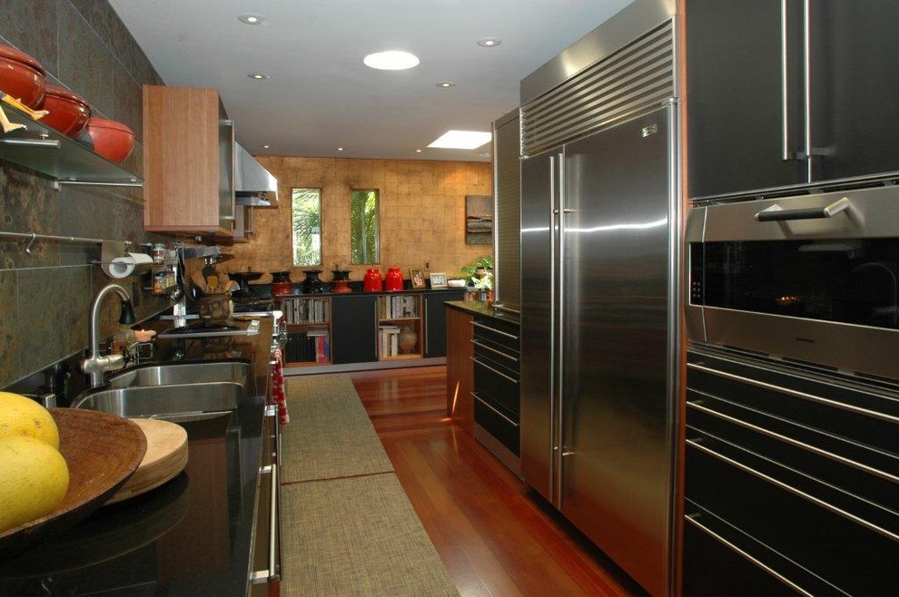 Large zen galley medium tone wood floor and brown floor enclosed kitchen photo in San Diego with a double-bowl sink, flat-panel cabinets, black cabinets, granite countertops, gray backsplash, stone tile backsplash, stainless steel appliances and no island
