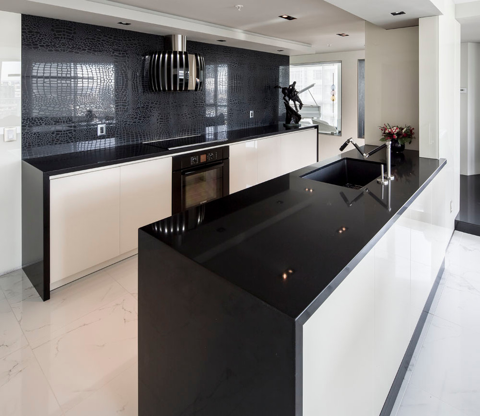 This is an example of a small modern galley open plan kitchen in Vancouver with flat-panel cabinets, engineered stone countertops, black splashback, an island, a submerged sink, beige cabinets, glass sheet splashback, stainless steel appliances, marble flooring and white floors.