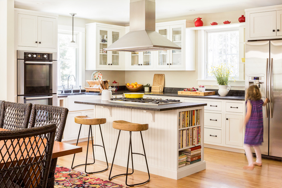 Mittelgroße Klassische Wohnküche in L-Form mit Landhausspüle, Glasfronten, weißen Schränken, Küchengeräten aus Edelstahl, hellem Holzboden, Kücheninsel, Granit-Arbeitsplatte, Rückwand-Fenster und braunem Boden in Portland Maine