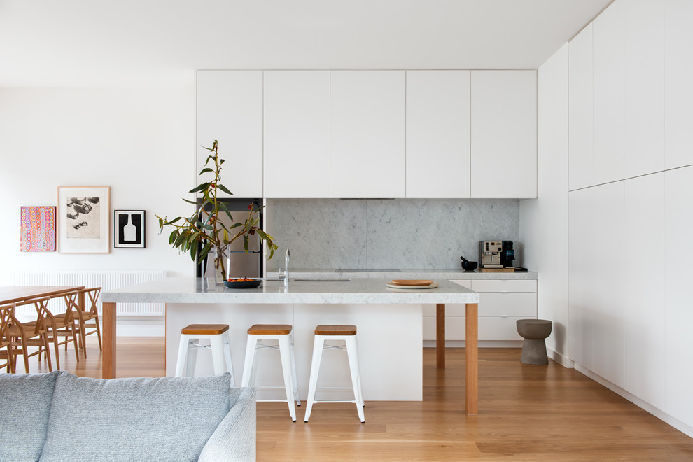Bild på ett funkis grå linjärt grått kök med öppen planlösning, med en dubbel diskho, släta luckor, vita skåp, marmorbänkskiva, grått stänkskydd, stänkskydd i marmor, rostfria vitvaror, ljust trägolv, en köksö och beiget golv