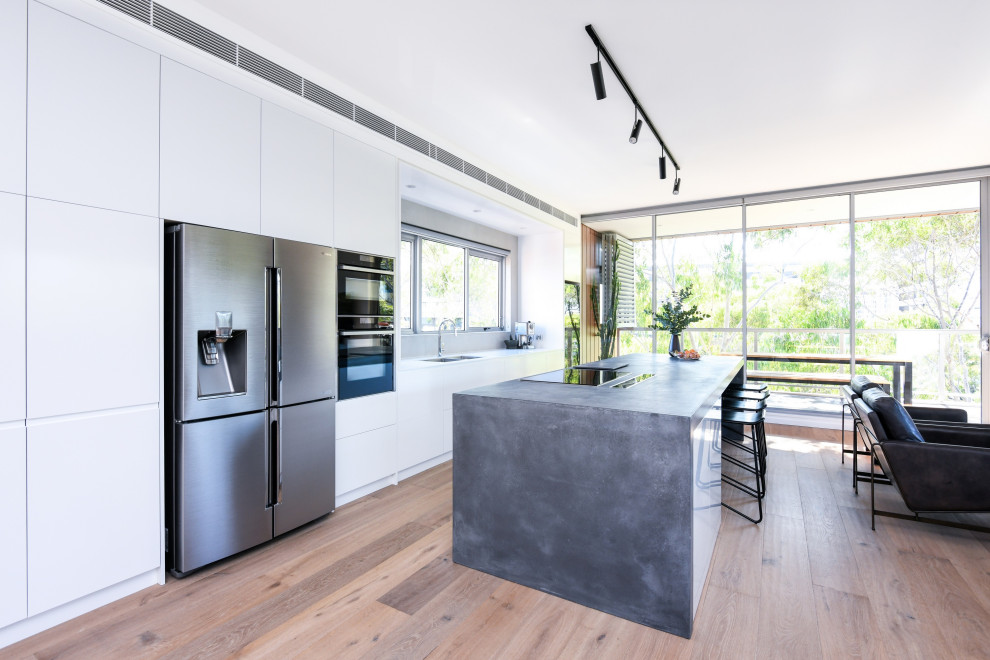 Inspiration for a large modern galley open plan kitchen in Sydney with a submerged sink, flat-panel cabinets, white cabinets, concrete worktops, grey splashback, black appliances, light hardwood flooring, an island, brown floors and grey worktops.