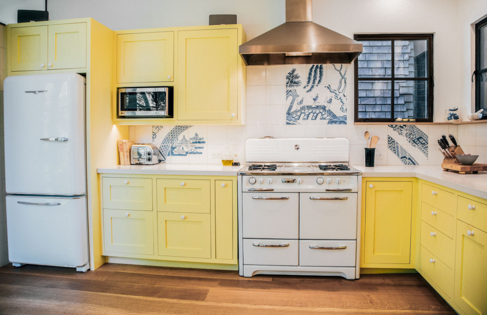 Inspiration for a large bohemian l-shaped kitchen pantry in San Francisco with a belfast sink, shaker cabinets, yellow cabinets, white splashback, porcelain splashback, white appliances, medium hardwood flooring, no island, brown floors and white worktops.