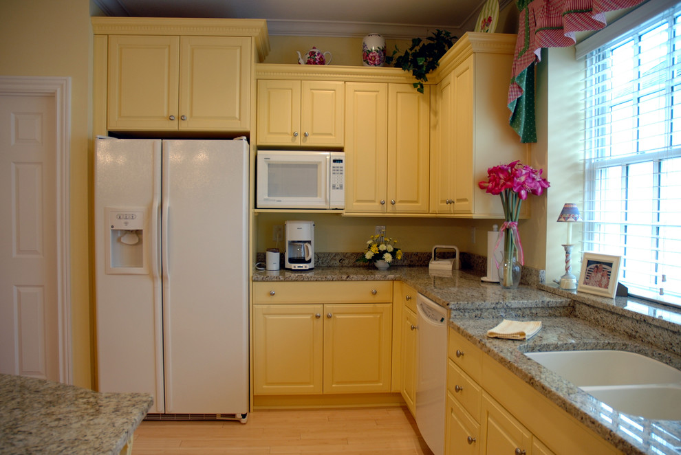 Modelo de cocina clásica con fregadero bajoencimera, armarios con paneles con relieve, puertas de armario amarillas, encimera de granito, electrodomésticos blancos, suelo de madera clara y una isla