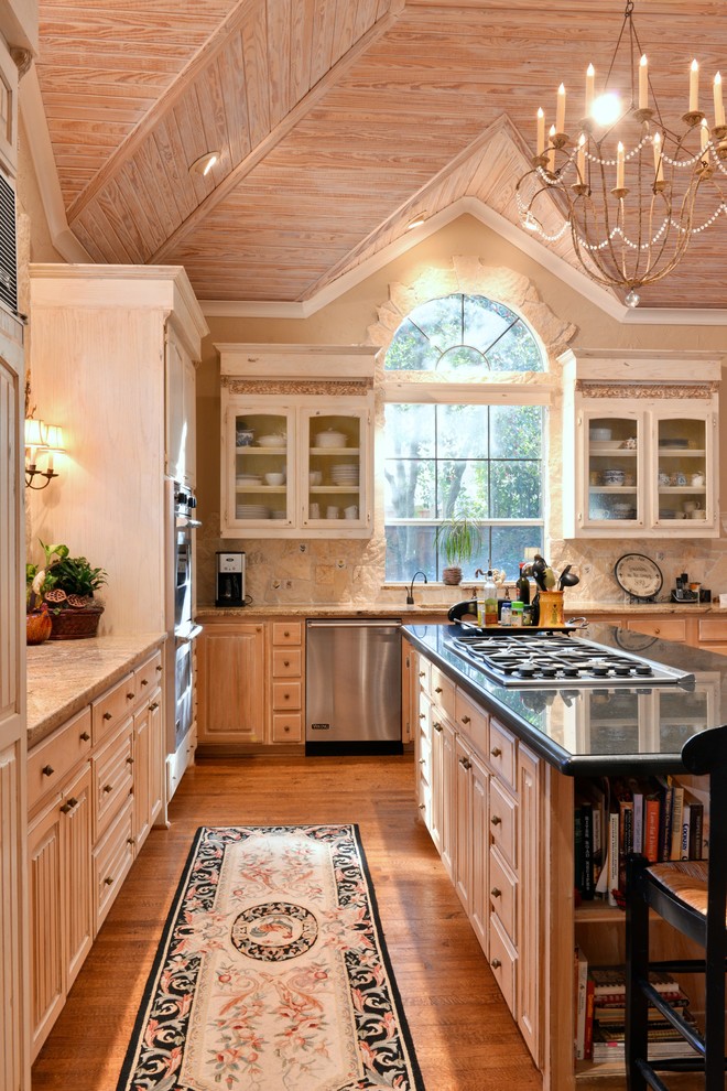 Example of a mountain style l-shaped medium tone wood floor kitchen design in Dallas with granite countertops, raised-panel cabinets, light wood cabinets, beige backsplash, stainless steel appliances, an island and limestone backsplash