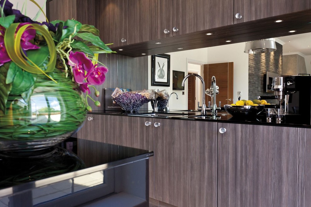 Photo of a large contemporary single-wall kitchen/diner in Hertfordshire with a built-in sink, flat-panel cabinets, dark wood cabinets, glass sheet splashback, black appliances and an island.